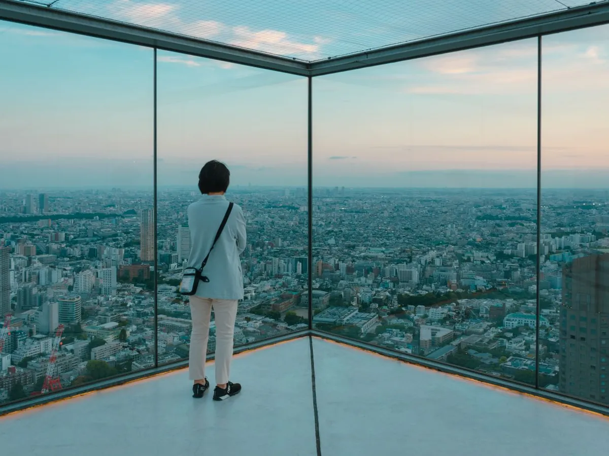 A visitor at the glass edge of the Shibuya Sky rooftop deck, looking over Tokyo at dusk
